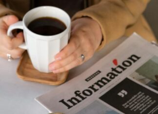 Osoba sedi za stolom na kojem se nalaze novine dok drži šolju kafe.