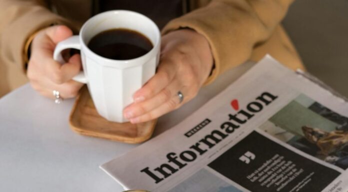 Osoba sedi za stolom na kojem se nalaze novine dok drži šolju kafe.