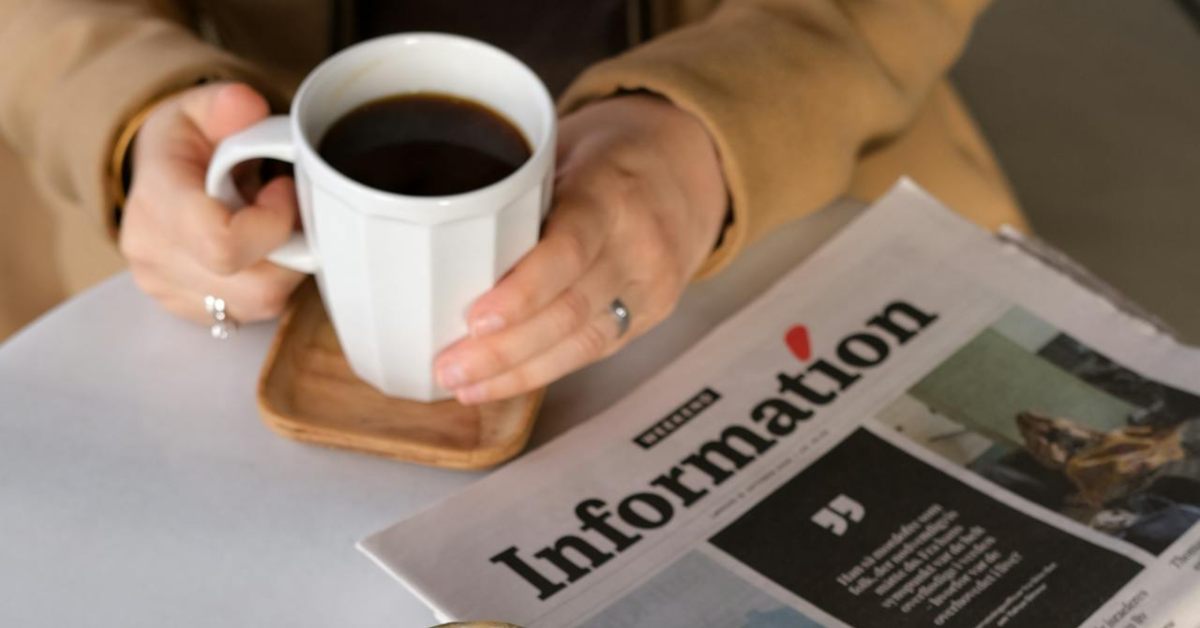 Osoba sedi za stolom na kojem se nalaze novine dok drži šolju kafe.