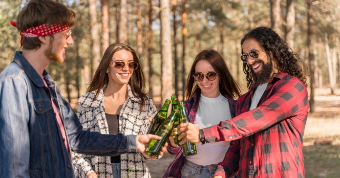 Dva momka i dve devojke nazdravljaju stojeći sa flašama piva u ruci u prirodi