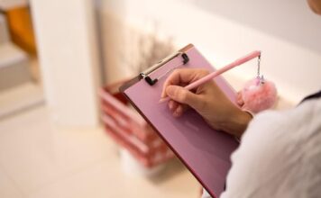 Ruka piše na ružičastoj tabli sa stezaljkom ružičastom olovkom sa plišanim priveskom.