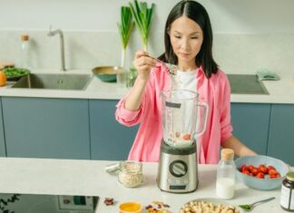 Žena u roze košulji dodaje ovsene pahuljice u blender na kuhinjskom pultu.