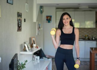 Kućni trening ili teretana – šta je bolje za početnike? Žena u crnom sportskom kompletu drži dve žute bučice u kuhinji.