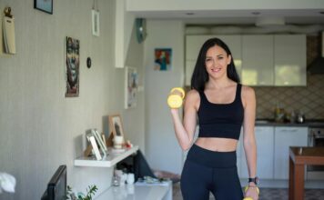 Žena u crnom sportskom kompletu drži dve žute bučice u kuhinji.