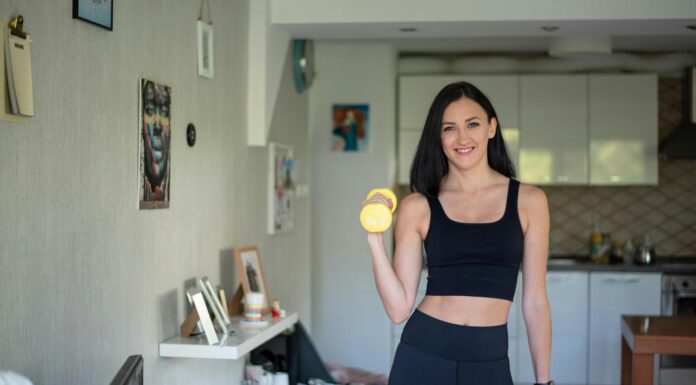 Kućni trening ili teretana – šta je bolje za početnike? Žena u crnom sportskom kompletu drži dve žute bučice u kuhinji.
