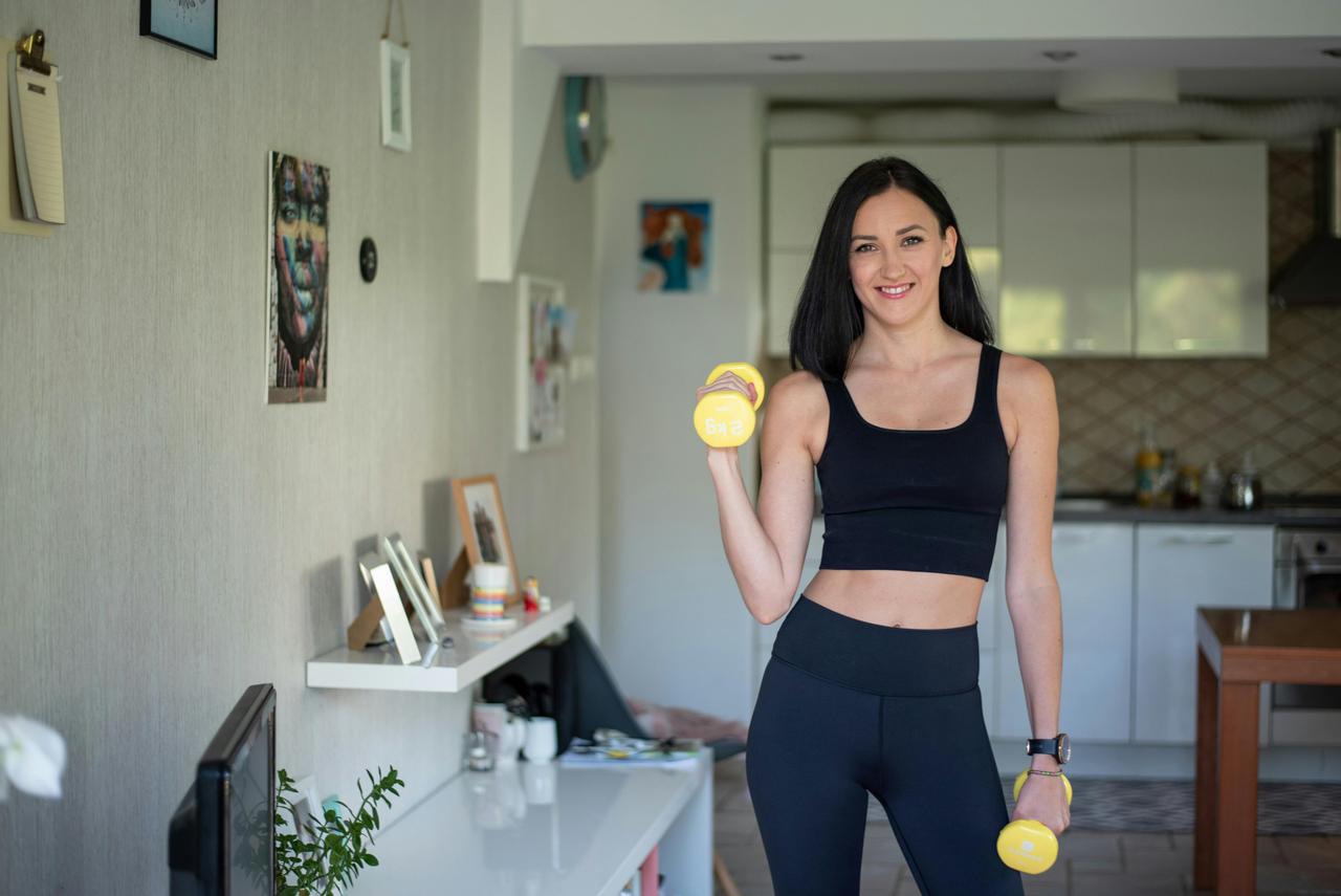 zena-sportski-bucice-kuhinja Žena u crnom sportskom kompletu drži dve žute bučice u kuhinji.